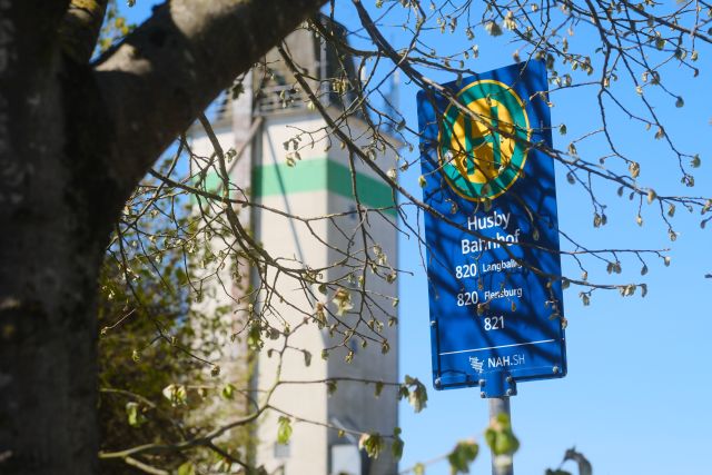 Bushaltestellenschild Husby Bahnhof vor Baum und blauem Himmel